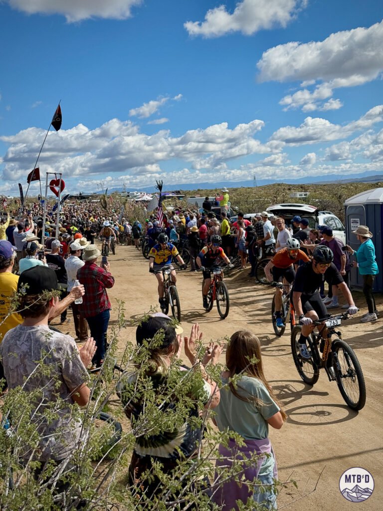 Riders launching from the start of 24 Hours in the Old Pueblo 2026 on the dirt road course at Willow Springs Ranch, surrounded by cheering spectators