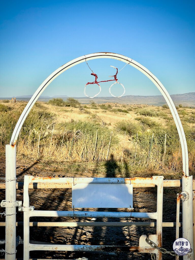 Handmade wire mountain bike figure hanging on gate at cattle trough in Honeybee Rail X Ranch trail network, Tucson Arizona