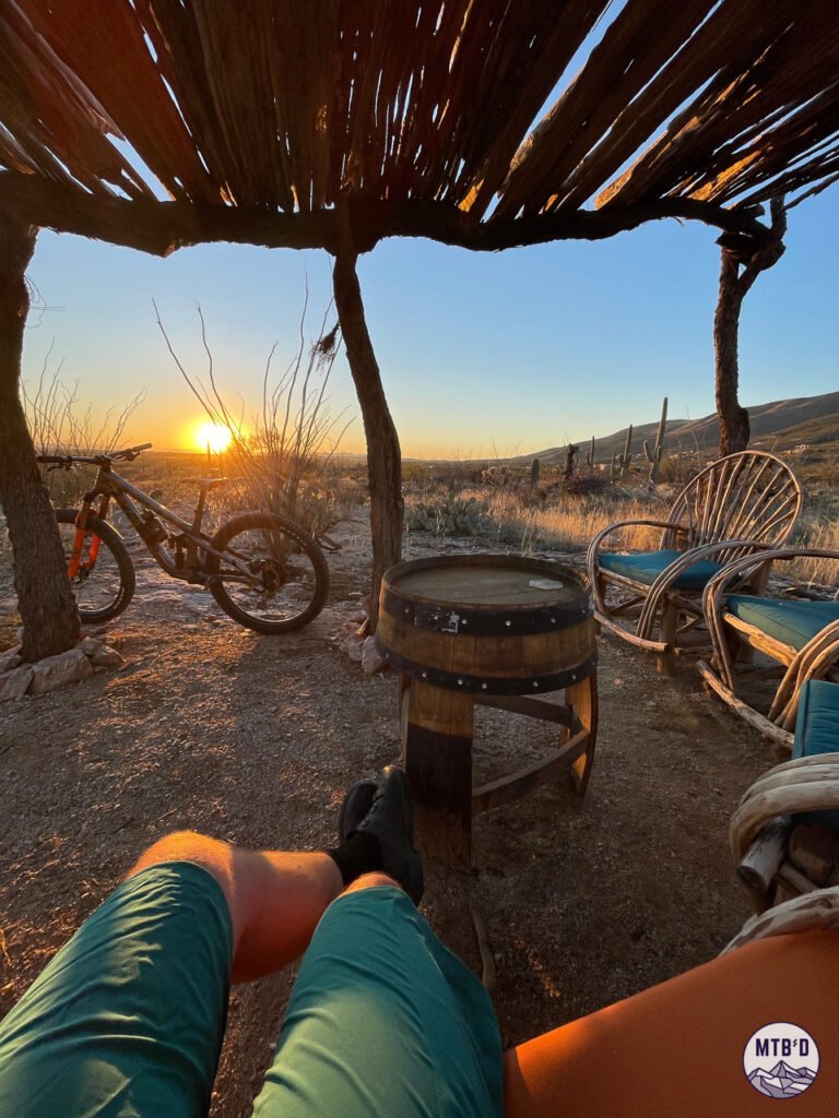 Mountain biker resting in trailside shade structure with feet extended and mountain bike in background at sunset near the bottom of La Milagrosa Trail, Tucson Arizona